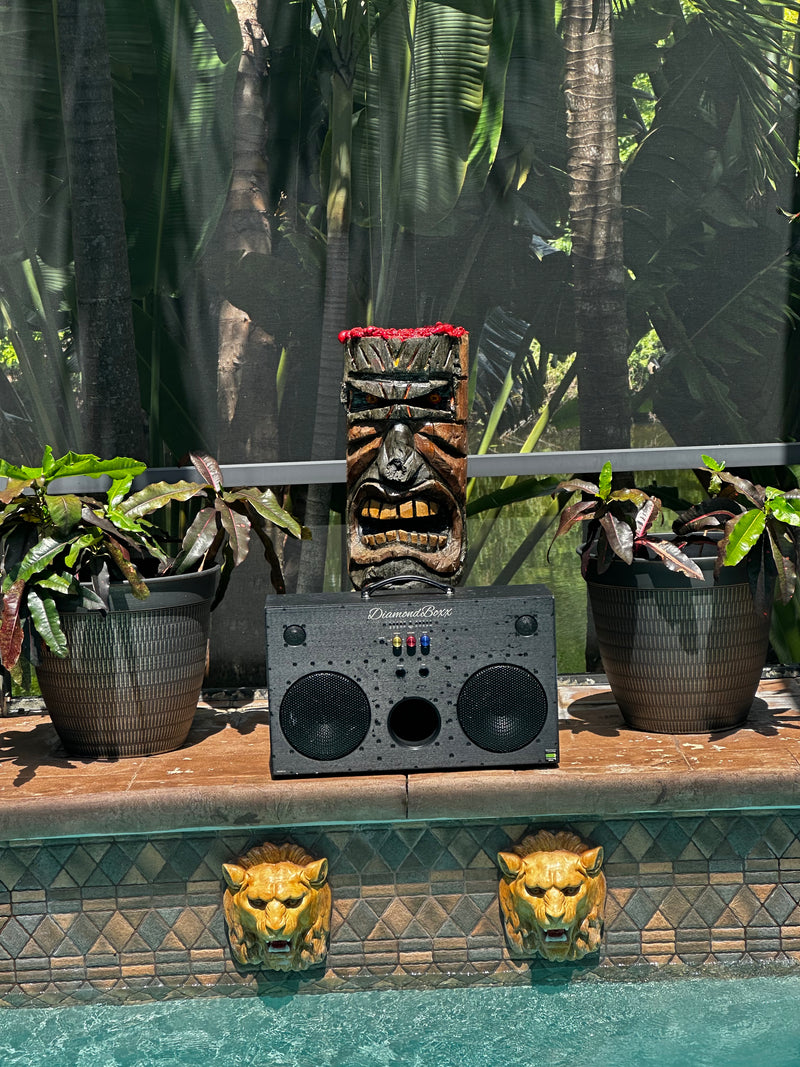 A black DiamondBoxx L3 speaker beside a pool with plants and a tiki sculpture.